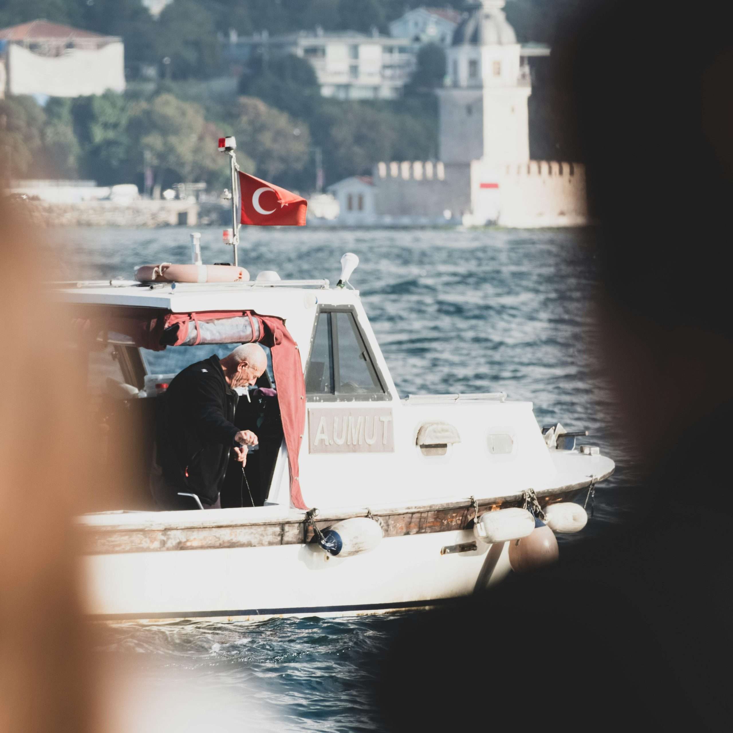 A man on a boat with the Turkish flag sails near a historic waterside tower.