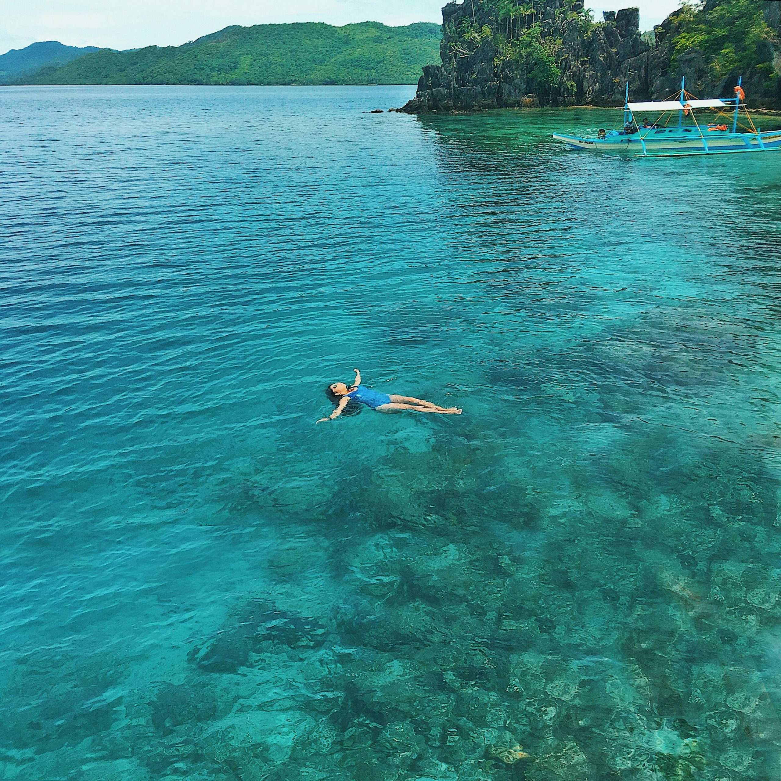 Relaxing scene of a woman floating in clear turquoise water in Palawan, Philippines, surrounded by lush greenery.
