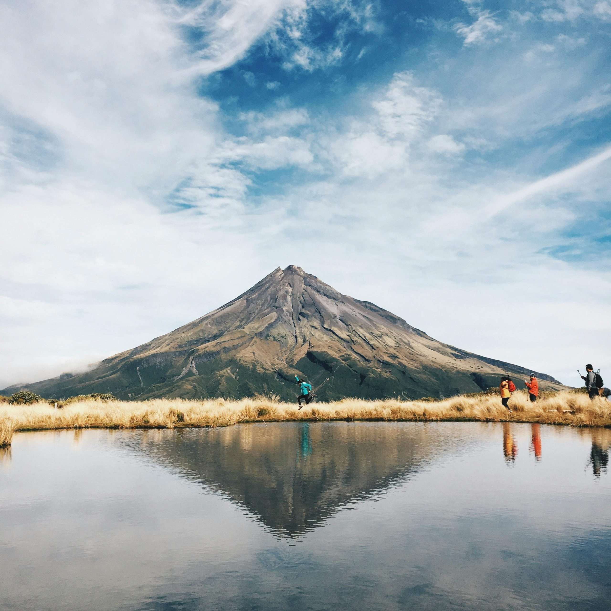 Hikers by a reflective lake with a stunning mountain backdrop on a clear day.