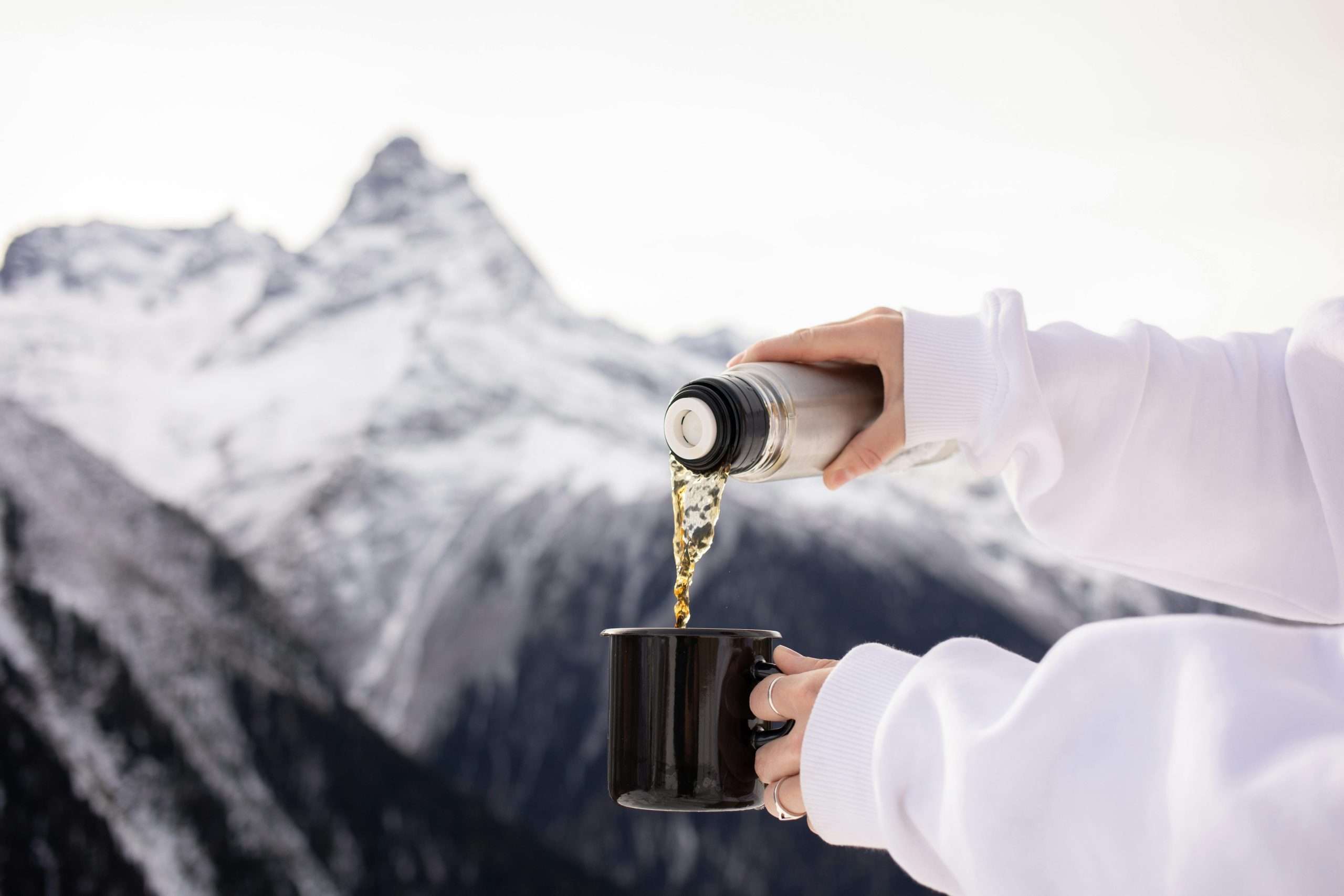 A hot beverage being poured into a mug against a snowy mountain backdrop.
